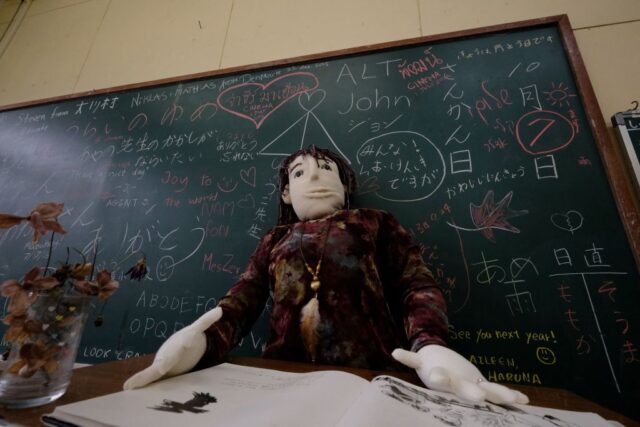 Doll sitting in front of a classroom blackboard with writing on it.